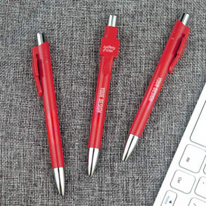 A set of three red personalised business pens with silver accents, resting on a textured grey surface near a keyboard.