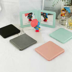 A set of square travel makeup mirrors in black, grey, pink, and mint green, arranged with a decorative figure.