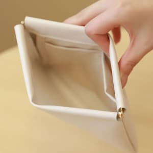 A hand holding a cream cosmetic bag with a clasp, featuring an interior pocket.
