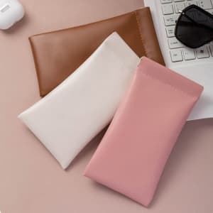 Three sunglasses storage bags in brown, white, and pink, placed on a surface near a keyboard and sunglasses.