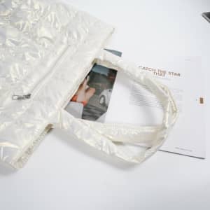 A large quilted tote bag in a glossy white finish, featuring handles and a zippered pocket, placed beside a magazine.