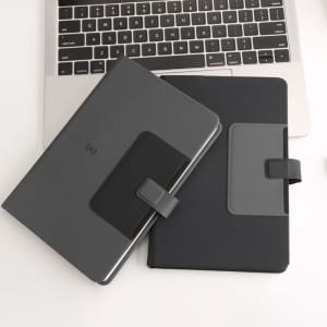 A grey notebook with a mobile phone holder, featuring a sleek design, rests on a desk alongside a laptop.