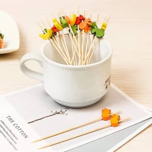 A white cup holds colourful disposable fruit cocktail picks featuring various fruit designs on wooden skewers.