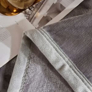 A soft grey flannel lap blanket with a lighter grey trim, laid on a surface alongside a drink and book.
