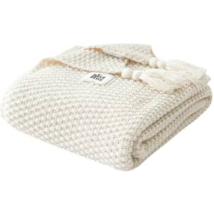 A knitted cream-coloured throw blanket featuring decorative tassels and a logo. Soft and textured design.