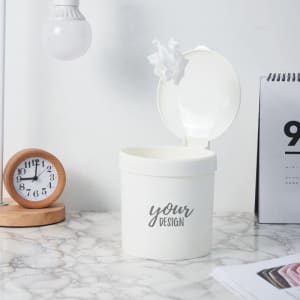 A white mini desktop trash can with a lid, placed on a marble surface, alongside a wooden clock and documents.