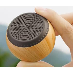 A portable wireless speaker with a wooden finish and a black speaker grille, held in a hand outdoors.