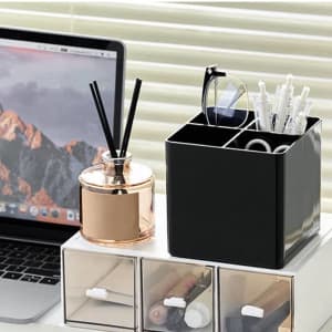 A black makeup brush holder organiser with multiple compartments, next to a gold candle holder and pens, on a workstation.