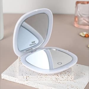 A white portable folding LED mirror with a magnification feature, resting on a textured surface.