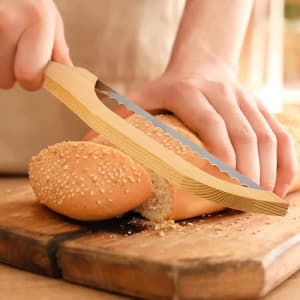 A serrated bread knife with a wooden handle slicing through a sesame seed roll on a wooden cutting board.