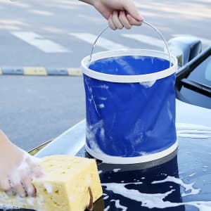 A foldable car washing bucket in blue with a handle, alongside a sponge and soapy water on a car's surface.