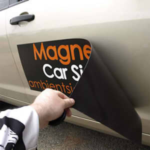 A hand holding a custom car magnet, showcasing a black backing and vibrant orange text, ready to be applied to a vehicle.