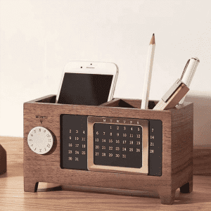 A wooden desk calendar with a pen holder, featuring a clock, a pencil, and space for a mobile phone.