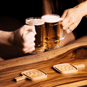 Two wooden pickleball-shaped coasters in a natural finish, placed on a wooden table with two beer mugs clinking above.