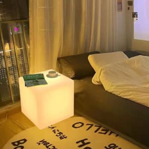 A glowing white cube serves as a bedside table next to a bed, complemented by a book and a cup.