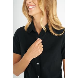 A black short sleeve shirt with a button-up front features a woman smiling while adjusting the collar.