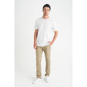 A man stands wearing a plain white t-shirt and beige pants, with one hand in his pocket.