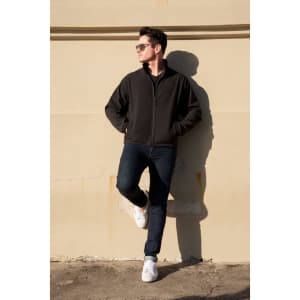 A soft shell jacket in black worn by a man leaning against a wall, paired with dark jeans and white sneakers.