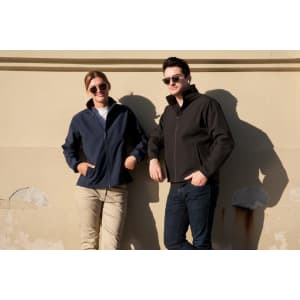 A soft shell jacket in navy worn by a woman and a black jacket worn by a man, both standing against a wall.