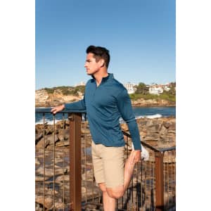 A man in a blue long-sleeve top and beige shorts stretches by the water, with rocky terrain in the background.