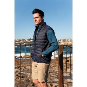 A man stands on rocky beachwearing a navy blue puffer vest and a teal long-sleeve top, with coastline in the background.