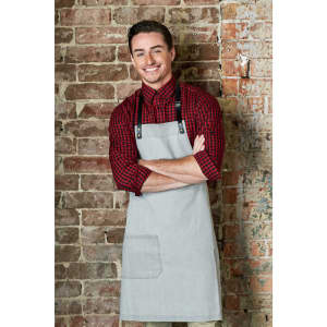 A grey bib apron with adjustable straps, worn over a red and black checkered shirt, against a brick wall background.