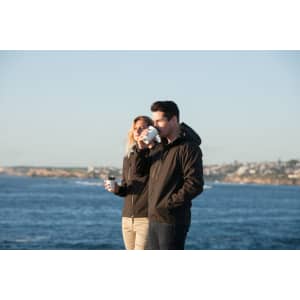 A couple wearing black soft shell jackets stands by the water, holding coffee cups and enjoying the view.