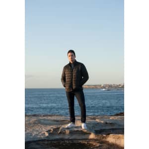 A man stands by the ocean wearing a black puffer jacket, blue jeans, and white shoes, with a coastal background.