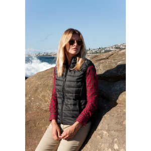 A black puffer vest worn by a woman with long blonde hair, sitting on a rock by the sea.