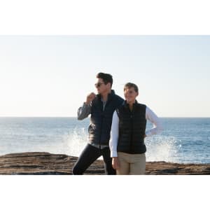 A navy puffer vest for ladies on a woman standing beside a man near the ocean, both wearing casual outfits.