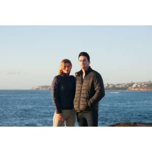 A black puffer jacket worn by a man and a navy puffer jacket worn by a woman against a coastal backdrop.