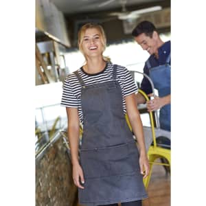 A woman in a dark grey bib apron over a striped shirt stands smiling in a café setting, with a man in the background.