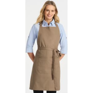A woman models a beige bib apron featuring a front pocket, paired with a blue shirt. The apron has a branding logo.