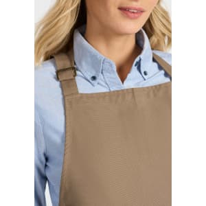 A brown bib apron is worn over a light blue shirt, featuring an adjustable strap and a subtle logo.