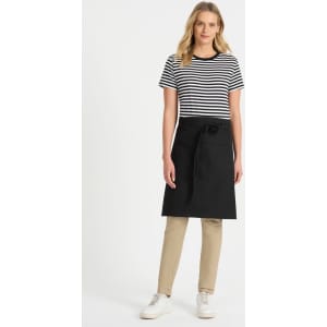 A woman wears a black and white striped t-shirt paired with a black apron and beige pants, standing against a plain background.