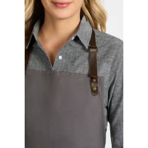 A ladies long sleeve button-up shirt in grey, featuring brown leather straps on an apron. It has a logo.