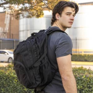 A black backpack is worn by a young man, featuring multiple pockets and a sturdy design, outdoors.