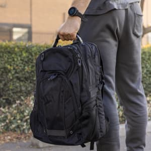 A black Eiger Backpack is held by a person wearing grey pants and a watch, set against a green hedge background.