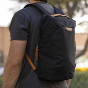 A person wearing a black backpack with an orange accent, featuring a logo, positioned against a blurred background.