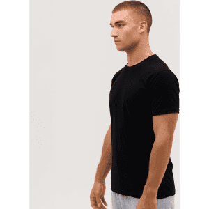 A black T-shirt is displayed on a model standing sideways, featuring a logo on the sleeve.