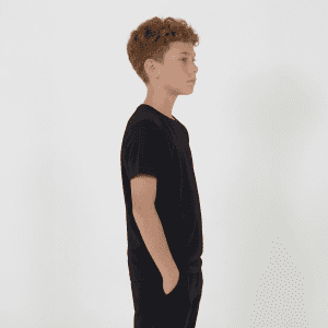 A black youth tee is displayed on a young person standing sideways with hands in pockets.