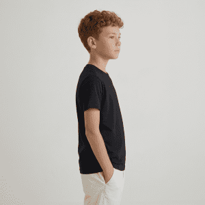 A child stands in profile wearing a black essential tee and white shorts, with hands in pockets.