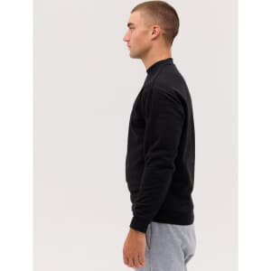 A black crewneck sweatshirt is worn by a model standing sideways, paired with grey sweatpants. The sweatshirt has a logo.