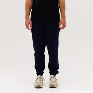 Navy track pants displayed on a person standing, paired with a black shirt and beige sneakers. The image features a logo.