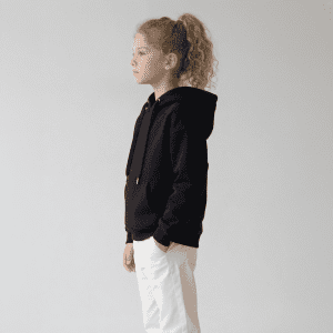 A black youth hoodie with a kangaroo pocket, drawstring hood, and a logo, paired with white trousers.