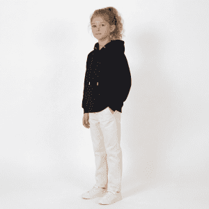 A young girl stands wearing a black hooded sweatshirt and white pants, with a light background. The hoodie has a logo.