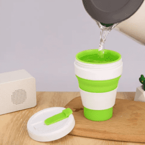 A collapsible silicone cup in green and white, being filled with water, beside a speaker on a wooden surface.