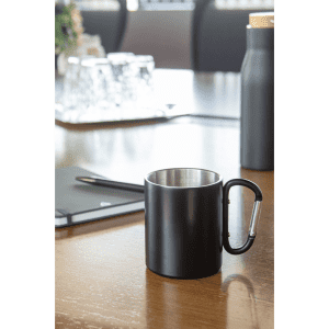 A black thermo mug with a silver interior and a carabiner handle on a wooden table, alongside a bottle and stationery.