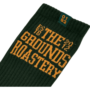 A green sock with yellow text featuring "THE GROUNDS ROASTERY" and a small logo at the top.