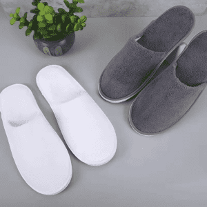 A pair of grey and a pair of white hotel slippers placed on a grey surface beside a small potted plant.